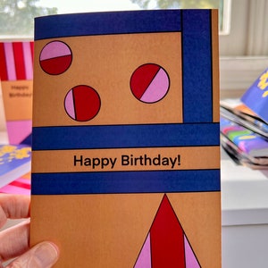 Puede incluir: Una tarjeta de cumpleaños con un diseño geométrico en rojo, rosa y azul sobre un fondo marrón. La tarjeta dice "¡Feliz cumpleaños!" en texto blanco.