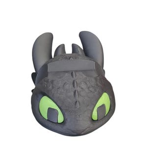 May include: A black and gray dragon head figurine with green eyes. The figurine has a textured surface and is a stylized representation of a dragon character. The dragon has large ears and a detailed face.