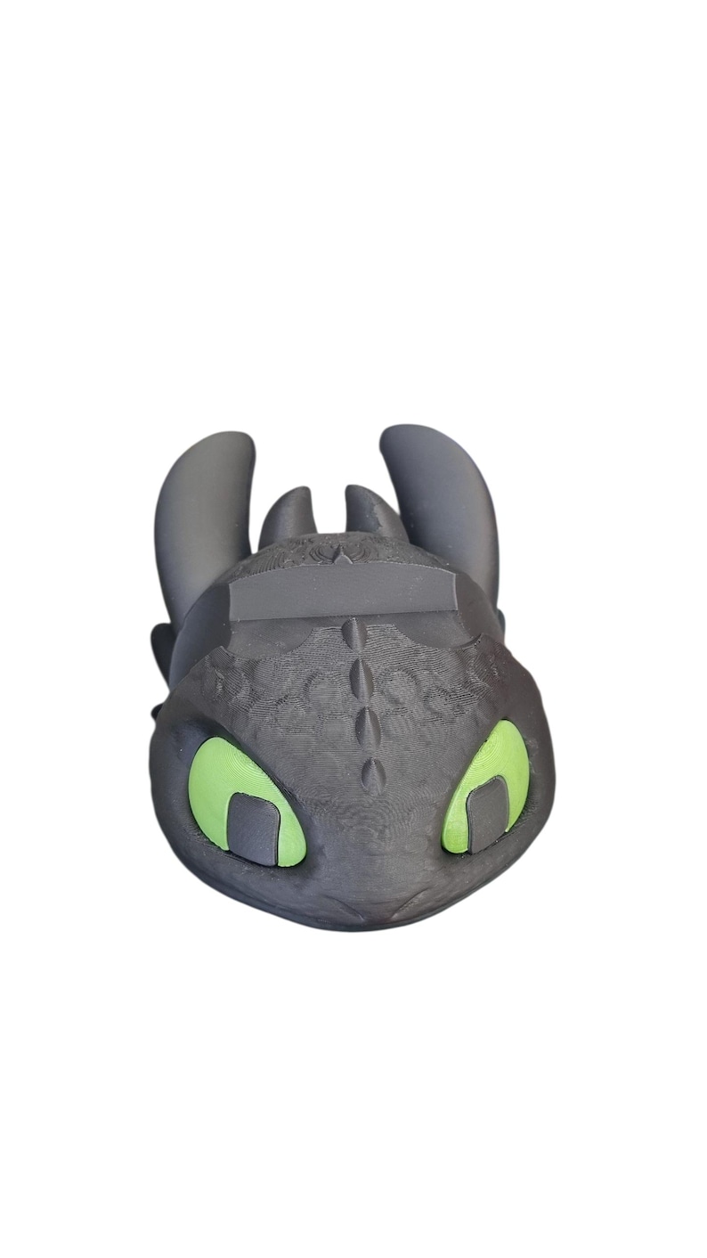 May include: A black and gray dragon head figurine with green eyes. The figurine has a textured surface and is a stylized representation of a dragon character. The dragon has large ears and a detailed face.