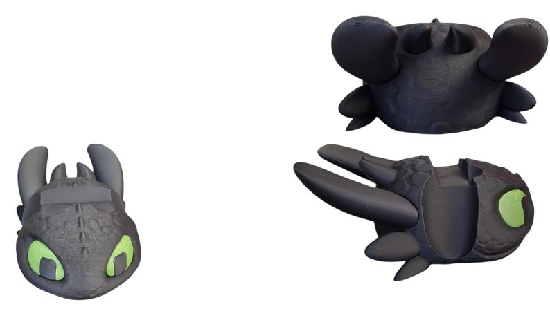 May include: Three black dragon-themed objects are displayed against a white background. The objects include a dragon head with green eyes, a dragon head with large ears, and a dragon wing. The objects are likely toys or collectibles.