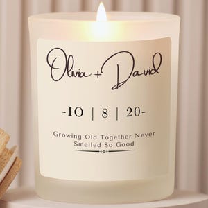 May include: A frosted glass candle with a lit wick and a cream-colored label. The label features the names Olivia + David, the date 10 | 8 | 20, and the text Growing Old Together Never Smelled So Good. A wooden lid sits to the left of the candle.