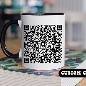 Op de afbeelding: Een witte keramische mok met een zwarte rand en handvat. Een zwart-witte QR-code is afgedrukt op de voorkant van de mok. De mok staat op een stapel boeken met de tekst "CUSTOM GIFT" gedrukt op het onderste boek.