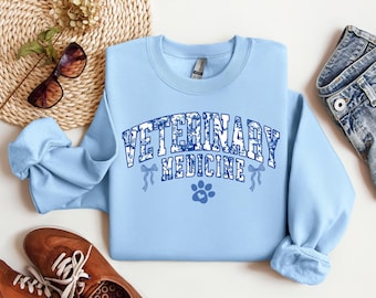 Könnte beinhalten: Hellblauer Pullover mit dem Text "Veterinary Medicine" in einer blauen und weißen gemusterten Schrift. Unter dem Text befindet sich ein Pfotenabdruck.
