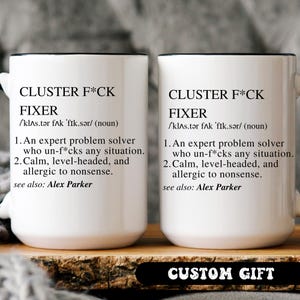 Op de afbeelding: Twee witte keramische mokken met zwarte randen en handvatten. Elke mok heeft de tekst "CLUSTER F*CK FIXER" met een definitie. De onderkant van de afbeelding heeft een zwarte banner met de tekst "CUSTOM GIFT".