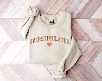 May include: A beige sweatshirt with the text "OVERStimulated" in colorful letters with a red heart in the middle.