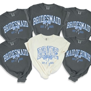 Puede incluir: Seis camisetas grises con texto azul y blanco y un lazo azul. Las camisetas dicen "Bride", "Maid of Honor" y "Bridesmaid" con el año "2025" impreso en cada una.