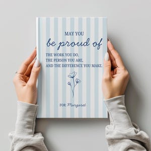 Może przedstawiać: Prostokątna książka z okładką w jasnoniebieskie i białe pionowe paski. Tekst brzmi: MAY YOU be proud of THE WORK YOU DO, THE PERSON YOU ARE, AND THE DIFFERENCE YOU MAKE. FOR: Margaret. Na środku znajduje się ciemnoniebieski rysunek kwiatka.