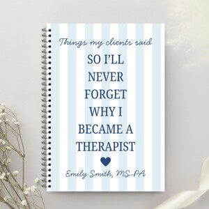 May include: A spiral-bound notebook with a light blue and white striped cover. The cover features the text "Things my clients said, SO I'LL NEVER FORGET WHY I BECAME A THERAPIST" in blue font. A blue heart is below the text, and the name "Emily Smith, MS-PA" is at the bottom.