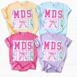May include: Four short-sleeved t-shirts in pastel colors: yellow, blue, pink, and purple. Each shirt features the letters "MDS" in pink, a pink bow, and floral and gingham patterns. The text "Hanna Smith RN" is also visible.