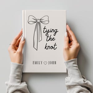 Puede incluir: Un libro blanco con una ilustración de lazo negro y la frase "tying the knot". Los nombres "Emily" y "John" están debajo, separados por un símbolo de corazón. El libro está sostenido por dos manos.