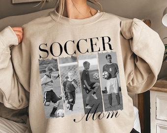 May include: A beige sweatshirt featuring the words "SOCCER Mom" in black. The word "SOCCER" is above four black and white images of soccer players. The sweatshirt is a neutral colour and has long sleeves.
