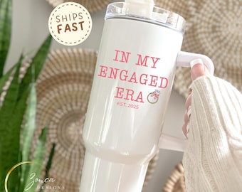 May include: White tumbler with a handle and a clear lid. The text "IN MY ENGAGED ERA" is printed in pink, along with a ring graphic and "EST. 2025". A "SHIPS FAST" sticker is visible.