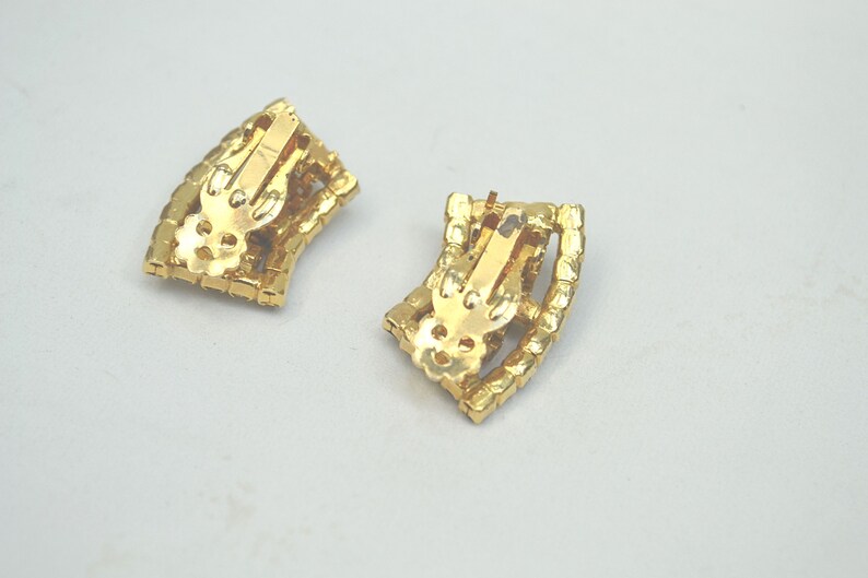 May include: A pair of gold tone clip-on earrings with a geometric design. The earrings are made of metal and have a shiny finish.