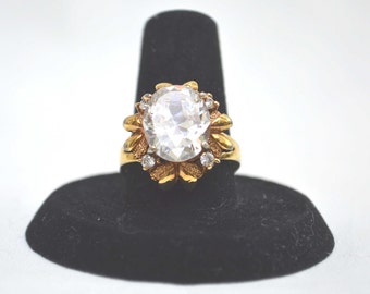 Op de afbeelding: Een gouden ring met een grote, heldere, ovale steen omringd door kleinere, heldere stenen. De ring ligt op een zwarte ringhouder.