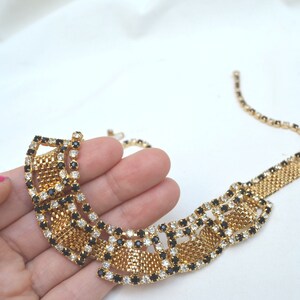 May include: A gold tone necklace with a geometric design featuring black and clear rhinestones. The necklace is made of a mesh chain and has a large, statement-making pendant.