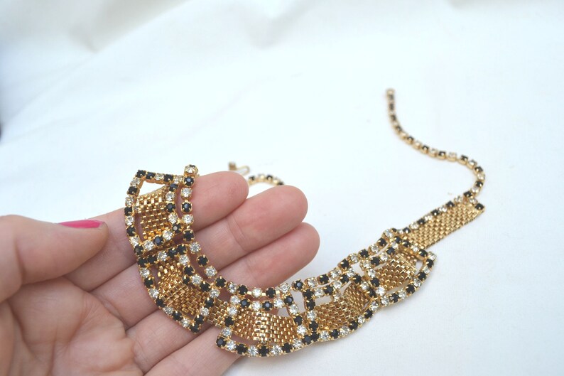 May include: A gold tone necklace with a geometric design featuring black and clear rhinestones. The necklace is made of a mesh chain and has a large, statement-making pendant.