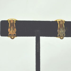 May include: A pair of gold-tone clip-on earrings with a round design. The earrings are on a black velvet display stand.