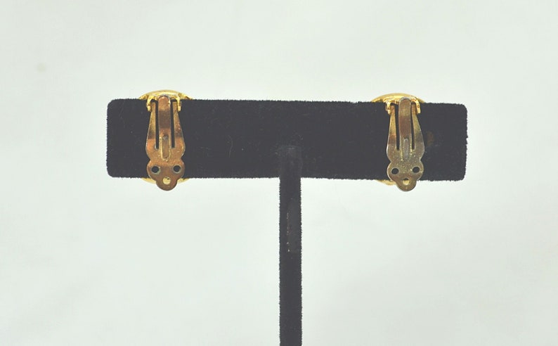 May include: A pair of gold-tone clip-on earrings with a round design. The earrings are on a black velvet display stand.