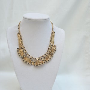 May include: A gold necklace with black and clear rhinestones in a geometric pattern. The necklace is displayed on a white mannequin bust.