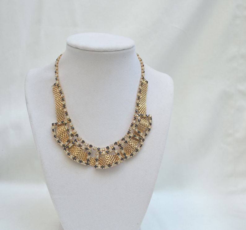 May include: A gold necklace with black and clear rhinestones in a geometric pattern. The necklace is displayed on a white mannequin bust.