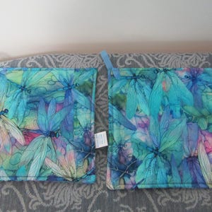 May include: Two blue and green pot holders with a dragonfly pattern. The pot holders have a light blue trim.