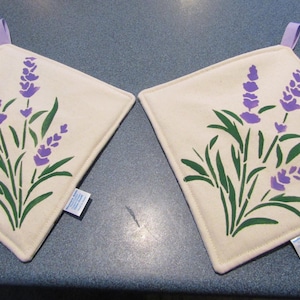 May include: Two square off-white pot holders with a lavender floral design. The lavender flowers are purple, and the stems and leaves are green. Each pot holder has a small purple loop for hanging.
