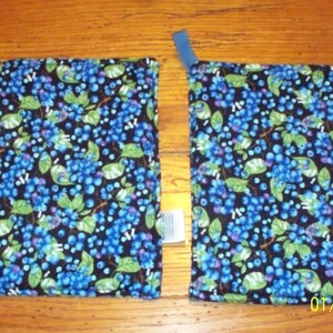 Op de afbeelding: Twee vierkante stoffen pannenlappen met een donkerblauwe achtergrond en een patroon van blauwe bessen en groene bladeren. Elke pannenlap heeft een kleine blauwe lus om op te hangen.