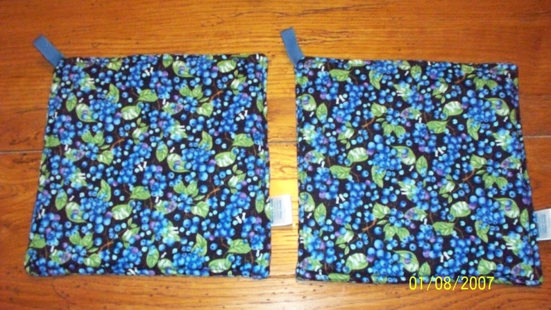 Op de afbeelding: Twee vierkante stoffen pannenlappen met een donkerblauwe achtergrond en een patroon van blauwe bessen en groene bladeren. Elke pannenlap heeft een kleine blauwe lus om op te hangen.