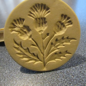 May include: A beige ceramic cookie cutter with a detailed design of three stylized thistles with leaves.