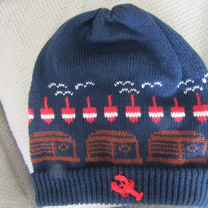 May include: Blue knitted beanie with a white, red, and brown pattern of buoys and lobster traps. A red lobster is attached to the front of the hat.