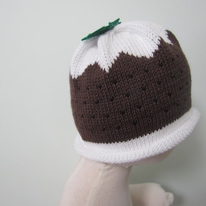 May include: A knitted hat in the shape of a Christmas pudding. The hat is brown with white accents and a green leaf on top. The hat has a white brim and is designed to look like a traditional holiday dessert.