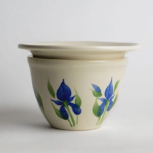 May include: Cream-colored ceramic butter dish with a lid. The dish is decorated with hand-painted blue iris flowers and green leaves. The design is simple and elegant, suitable for kitchen decor.