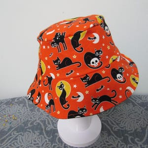 May include: Orange Halloween bucket hat with a repeating pattern of black cats, bats, moons, stars, and skulls. The hat is made of fabric and has a rounded crown and a wide brim. The hat is displayed on a white stand.