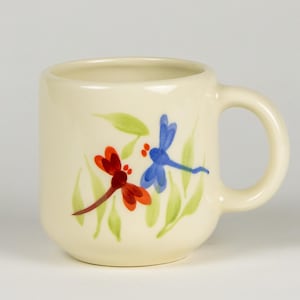 Può includere: Una tazza in ceramica color crema con un designo di libellula blu e rossa. La libellula è dipinta su un ramo verde con foglie.