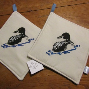 以下が含まれることがあります： 青と黒の loon のデザインが施された白い綿製の鍋つかみが2つ。鍋つかみには、吊り下げ用の青いループが付いています。一方の鍋つかみには、「Sierra's Attic」というテキストのタグが付いています。