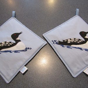 以下が含まれることがあります： 2つの正方形の、薄い青色の鍋つかみ。各鍋つかみには、赤い目をした黒と白のアビが、青い水面に浮かんでいるデザインが施されています。吊り下げ用の薄い青色のループが付いています。