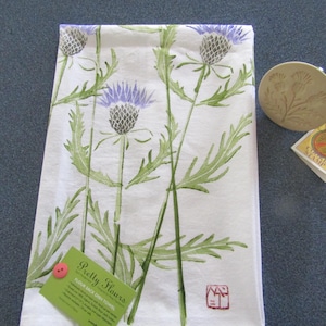 May include: White tea towel with a green and purple floral design. The design features multiple stylized thistle flowers. The text "Pretty Flowers" is printed on a green tag attached to the towel.