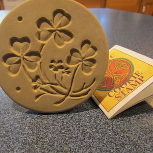 May include: A round, tan-colored cookie stamp with a raised shamrock design. The stamp has three large shamrocks and several smaller ones. A folded tag with the words "COOKIE STAMP" is next to the stamp.