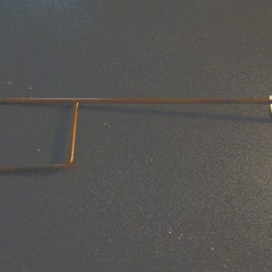 Pu&ograve; includere: Un oggetto metallico color rame con una forma rettangolare e una lunga barra dritta che si estende da esso. L'oggetto &egrave; posto su uno sfondo grigio scuro. Il design sembra essere uno strumento o un componente.