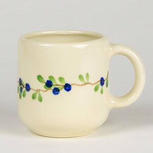 Può includere: Una tazza in ceramica bianca con un design a vite di bacche blu e verdi. La tazza ha una forma arrotondata e un manico.