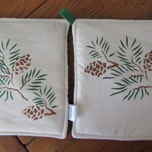 以下が含まれることがあります： 緑色の松の枝のステンシルデザインが施された白い綿製の鍋つかみが2つ。松の枝には茶色の松ぼっくりがついています。