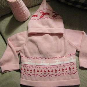 Può includere: Un maglione rosa lavorato a maglia con un cappuccio rosa abbinato. Il maglione presenta un motivo bianco e rosso di barche e aragoste.