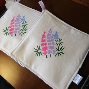 May include: Two white cotton pot holders with a blue trim. Each pot holder features a stenciled design of pink and purple lupine flowers with green leaves.