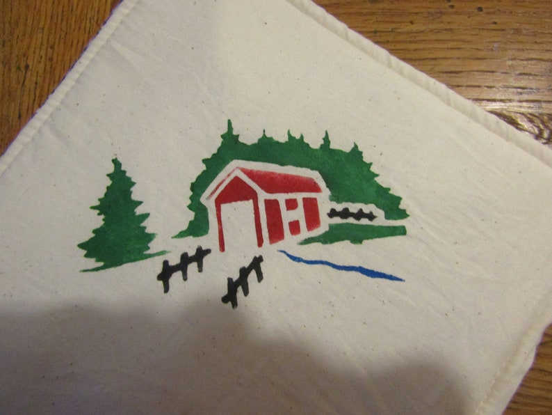 Op de afbeelding: Een witte doek met een handgeschilderd ontwerp met een rode overdekte brug, groene bomen, een blauwe beek en een zwart hek. Het ontwerp is gecentreerd op de doek.
