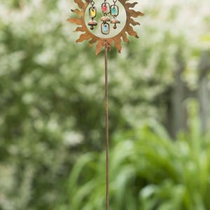 Pu&ograve; includere: Un paletto da giardino in metallo arrugginito con un design a forma di sole e ornamenti in vetro colorato. Il sole ha un centro circolare con ornamenti in vetro sospesi gialli, rossi e blu. Il paletto &egrave; in metallo scuro ed &egrave; in un giardino.