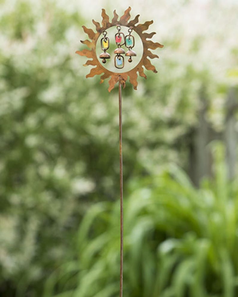 Pu&ograve; includere: Un paletto da giardino in metallo arrugginito con un design a forma di sole e ornamenti in vetro colorato. Il sole ha un centro circolare con ornamenti in vetro sospesi gialli, rossi e blu. Il paletto &egrave; in metallo scuro ed &egrave; in un giardino.