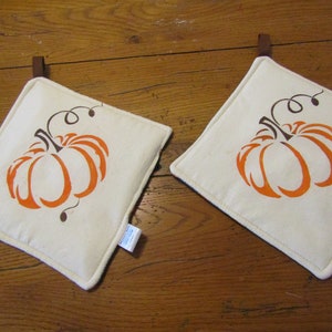 Op de afbeelding: Twee witte katoenen ovenwanten met oranje pompoenontwerpen. De pompoenen hebben bruine stelen en ranken.