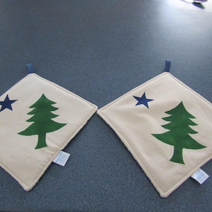 May include: Two square, off-white pot holders with a green tree and a blue star design. Each pot holder has a blue loop for hanging and a white border. The pot holders are approximately 8 inches square.