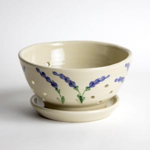 May include: Cream-colored ceramic colander with a matching saucer. The colander features hand-painted lavender sprigs in purple and green. It has several small holes for draining and is ideal for kitchen use.