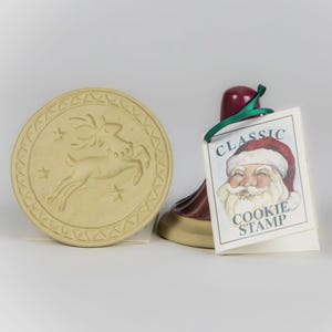 May include: A beige, round cookie stamp with a reindeer design and star accents. A red and gold cookie stamp with a Santa Claus image and the words "CLASSIC COOKIE STAMP" on a tag.